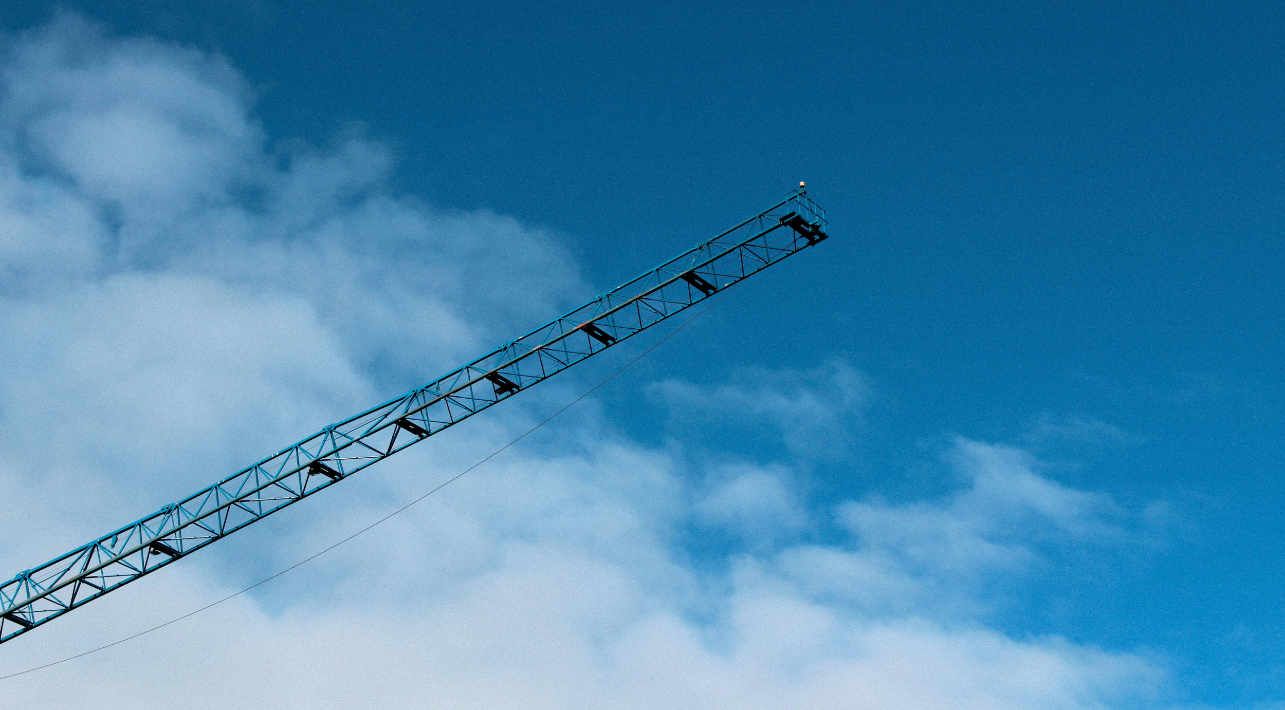 The top section of a blue construction crane extending diagonally into a bright blue sky with scattered white clouds.