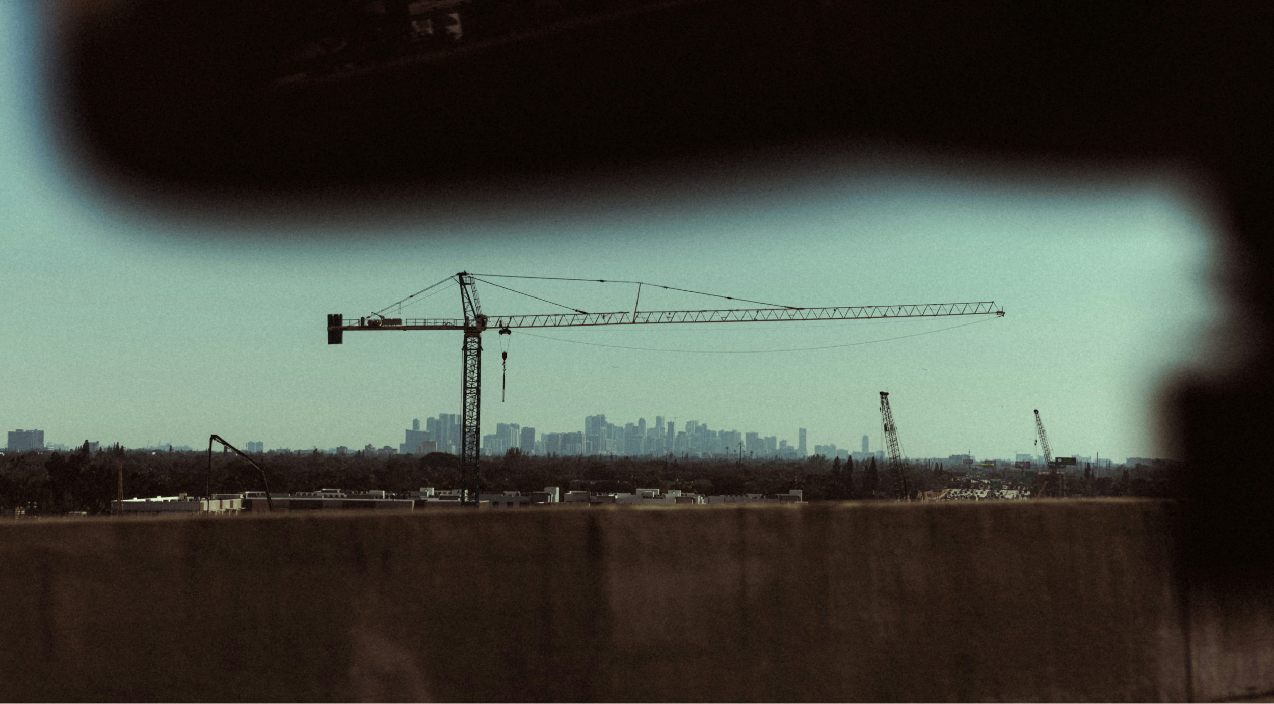 A large construction crane set against the skyline of a distant city, framed by the edges of a car window.
