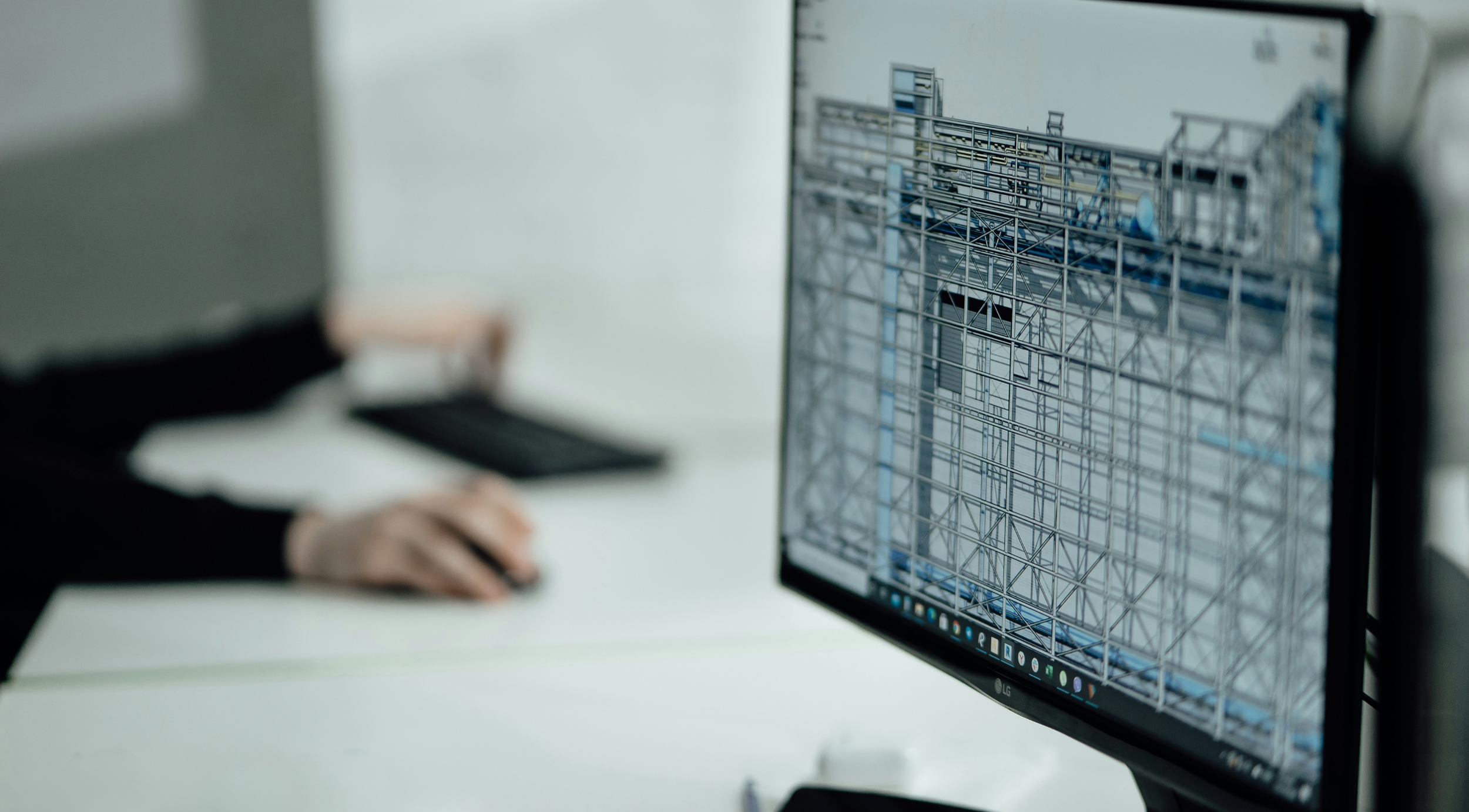 Person working at a desk on a computer displaying a detailed 3D architectural or structural design model, with focus on the monitor showing technical drawings.
